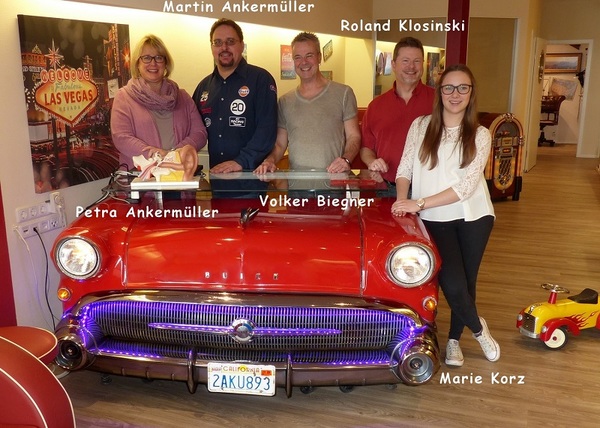 Fünf Personen stehen lächelnd hinter einem roten Oldtimer-Schreibtisch im Retro-Büro.